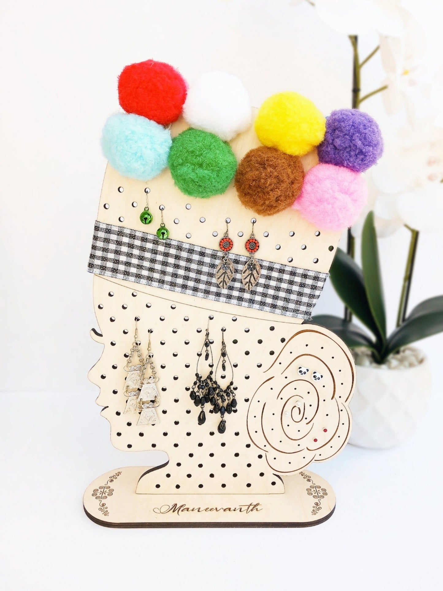This earring holder stand is in the shape of a woman's face wearing a traditional Hmong pom-pom hat and held by the base. Both pieces are made from Baltic Birch plywood and inspired by the Hmong pom-pom hat. The face holder is embellished with colorful 2 inch pom-poms and a checkered pattern ribbon accented with glitter. This earring holder has 127 big-sized holes and 42 small-sized holes to organize fish hook and stud earrings. A name and or word can be personalized on both ends of the base.
